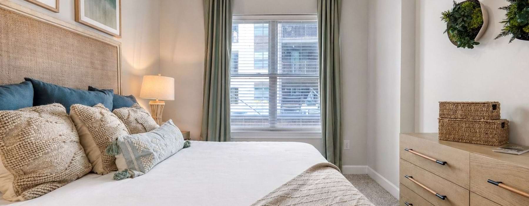 Bright apartment bedroom with a plush bed layered in neutral and blue accent pillows, framed artwork above the headboard, a wood dresser, soft carpeting, and a sunlit window with drapery at The Carson apartments in Charlotte, NC for rent.